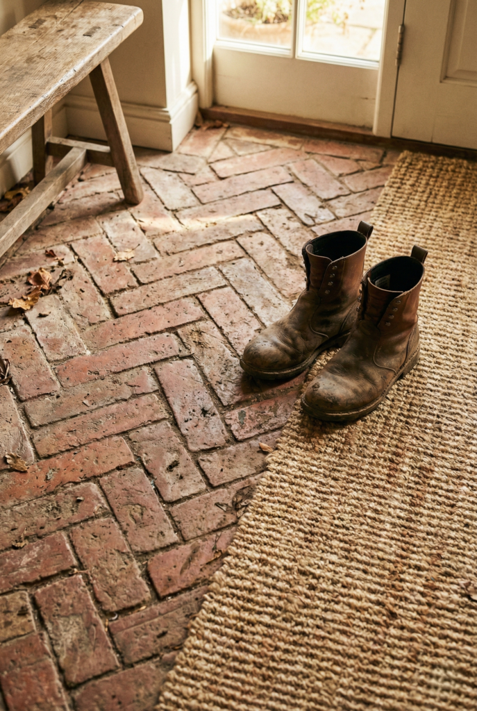 Close up of tumbled brick flooring in a cottage mudroom with leather boots and a woven rug.